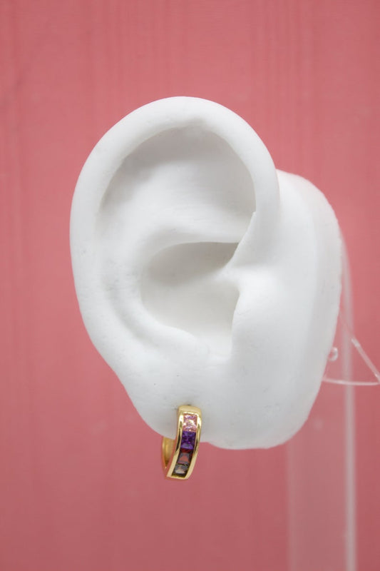 A white mannequin ear model displays a gold huggie hoop earring embedded with colorful gemstones against a pink background.