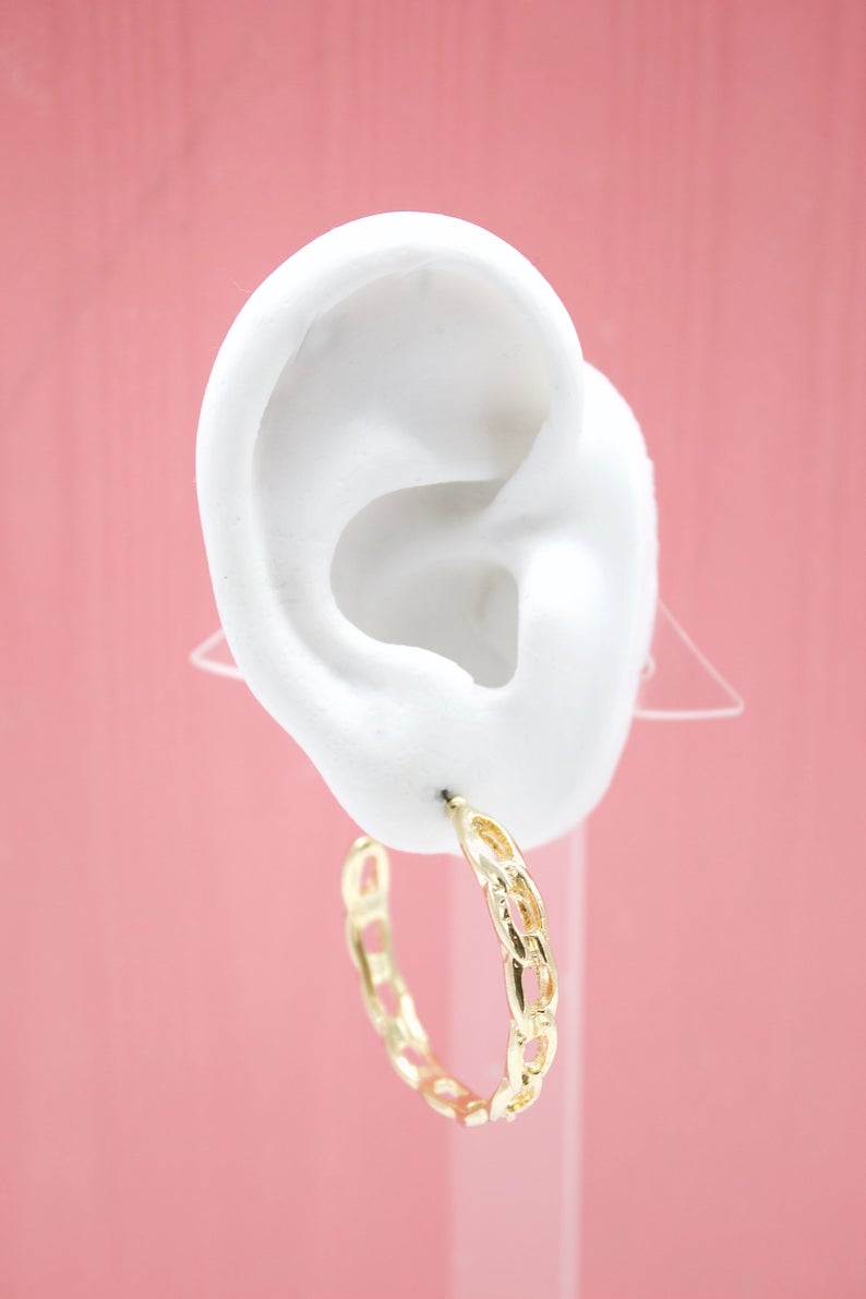 A white mannequin ear displays a stylish gold chain-link hoop earring against a pink background.