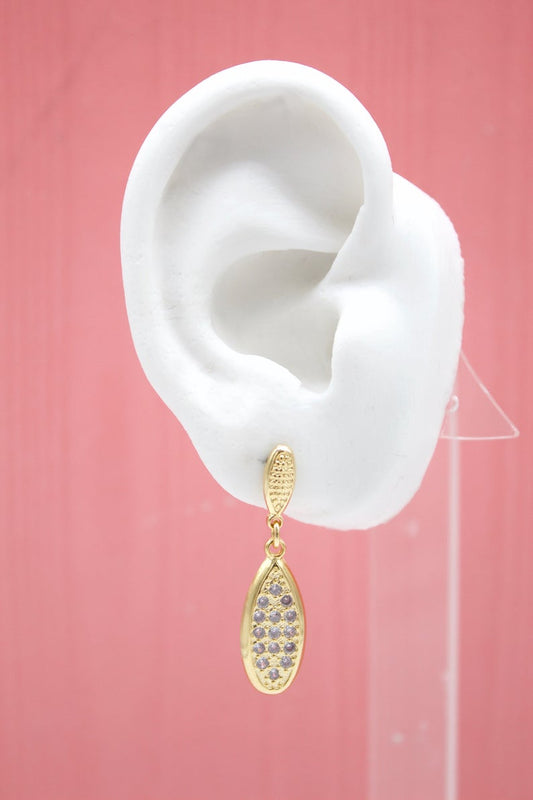 A white mannequin ear displays a gold teardrop pendant earring featuring purple pavé stones against a pink background.