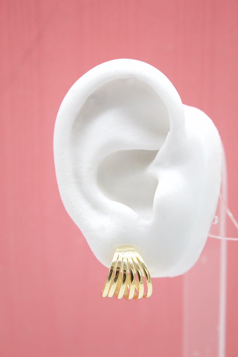 A white display ear model showcases a shiny gold multi-band huggie hoop earring against a soft pink background.