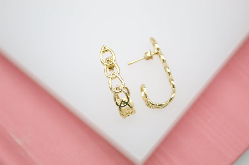 A pair of gold huggie hoop earrings featuring an interconnected heart pattern rests against a white and pink background.