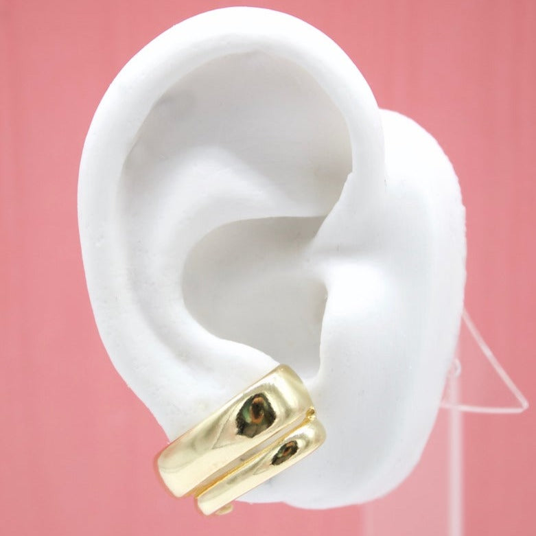 A white mannequin ear displays a polished, thick gold hoop earring against a soft pink background.