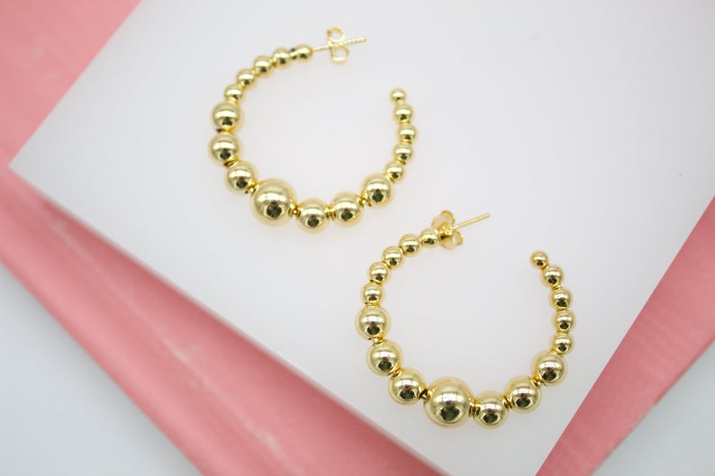 A pair of shiny gold ball hoop earrings rests on a white display surface.