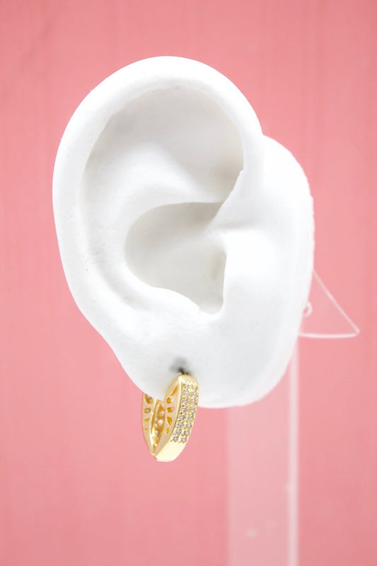 A white mannequin ear displays a sparkling gold huggie hoop earring encrusted with tiny cubic zirconia stones against a solid pink backdrop.
