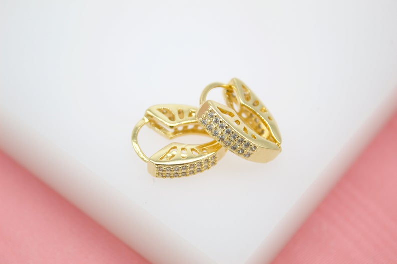 A pair of elegant gold huggie hoop earrings featuring sparkling pave-set cubic zirconia stones rests on a white display surface.