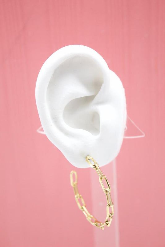 A white mannequin ear displays a modern gold paperclip chain hoop earring against a soft pink background.