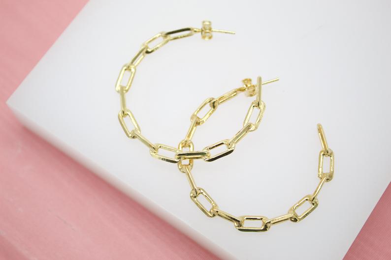 Two gold paperclip chain hoop earrings rest on a white surface against a pink background.