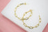 Two gold paperclip chain hoop earrings rest on a white surface against a pink background.