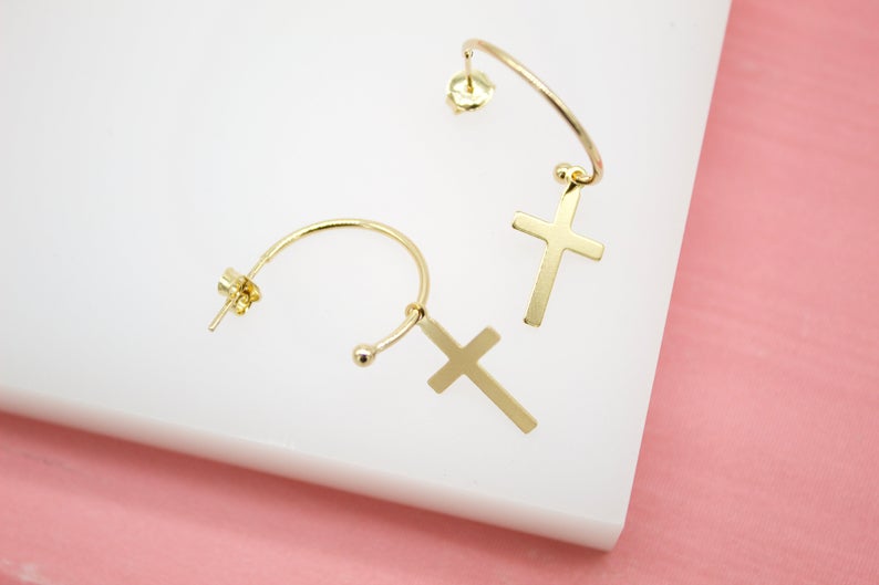 A pair of delicate gold cross hoop earrings rests on a white display against a pink background.