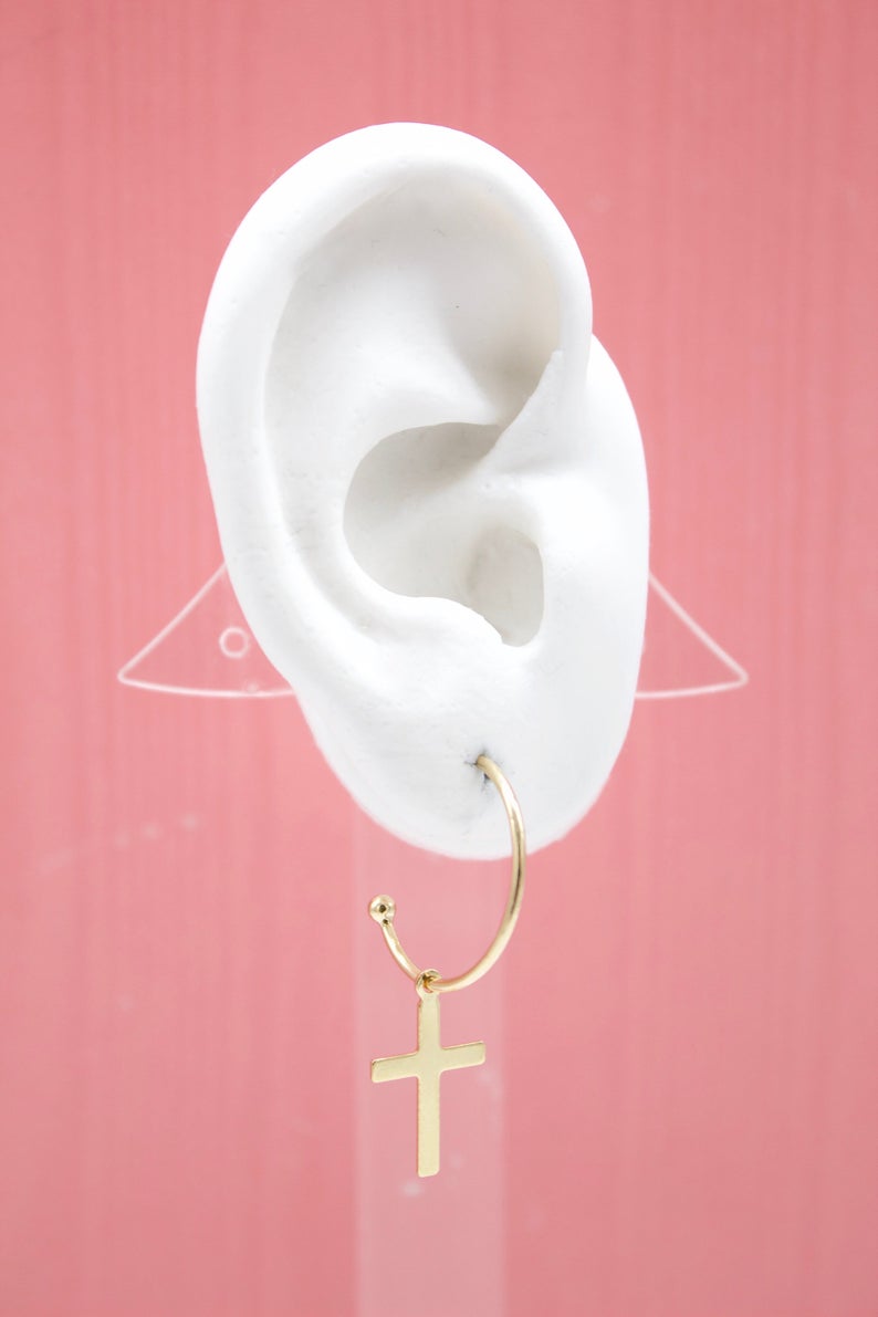 A white ear model displays a delicate gold hoop earring featuring a dangling cross charm against a solid pink background.