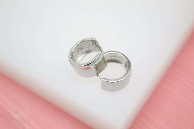 A pair of sleek silver huggie hoop earrings rests delicately on a white surface against a soft pink background.