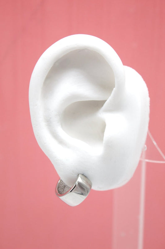 A white mannequin ear displays a chunky silver hoop earring against a solid pink backdrop.
