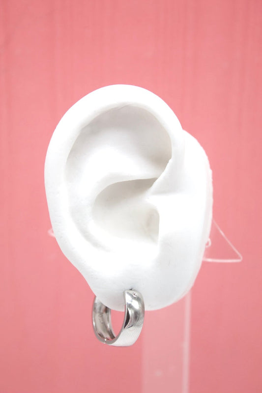 A white mannequin ear displays a polished silver huggie hoop earring against a pink background.