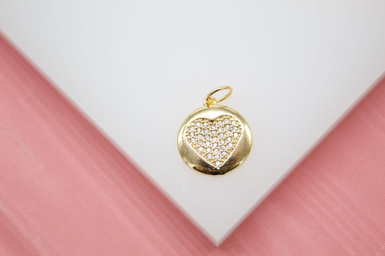 A gold medallion pendant features a pavé-set diamond heart on a white surface with a pink background.