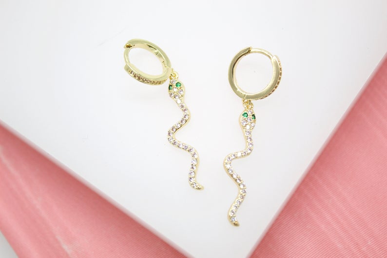 A pair of gold snake hoop earrings with pavé crystals and green eyes rests on a white surface.