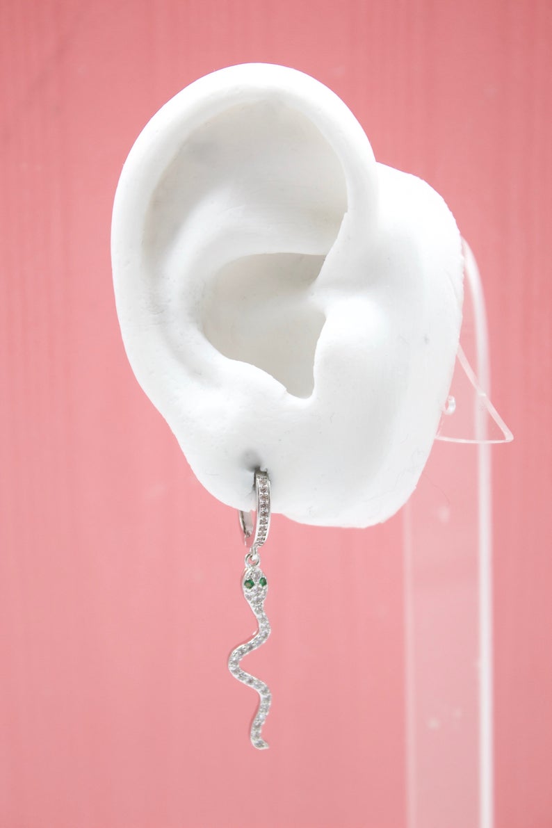 A white mannequin ear displays a sparkling silver snake huggie hoop earring with tiny green gemstone eyes against a solid pink background.