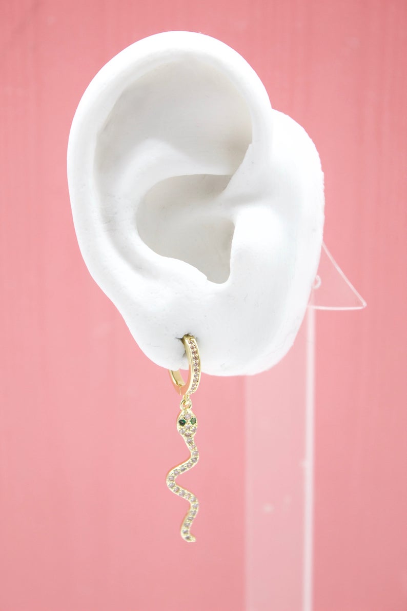 A white mannequin ear displays a gold snake huggie hoop earring adorned with sparkling pavé crystals and green gemstone eyes against a soft pink background.