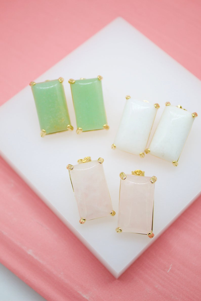 Three pairs of rectangular gemstone stud earrings in green, white, and pink, set in gold prongs, rest on a white surface against a pink background.
