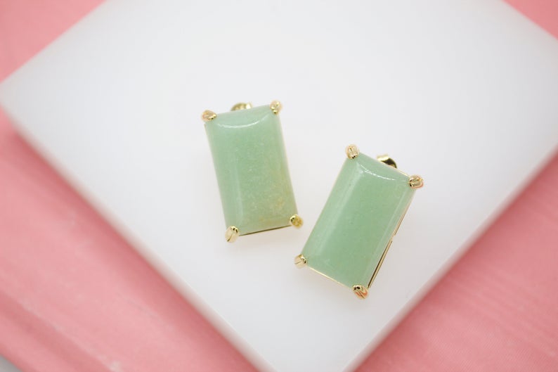 A pair of rectangular green aventurine stone stud earrings with gold settings rests on a white display against a pink background.