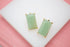 A pair of rectangular green aventurine stone stud earrings with gold settings rests on a white display against a pink background.