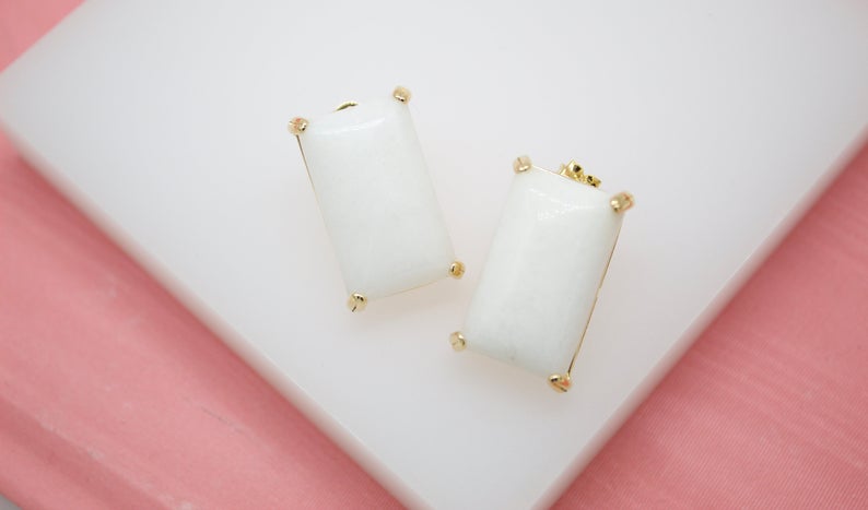 A pair of rectangular white stone earrings with gold prongs rests on a white display stand against a pink background.