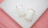 A pair of rectangular white stone earrings with gold prongs rests on a white display stand against a pink background.