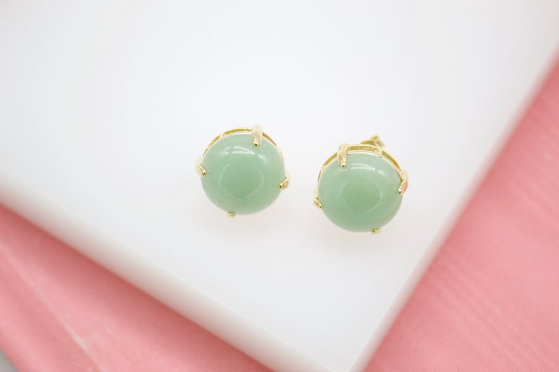 A pair of round green jade gemstone stud earrings in gold settings rests on a white display surface.