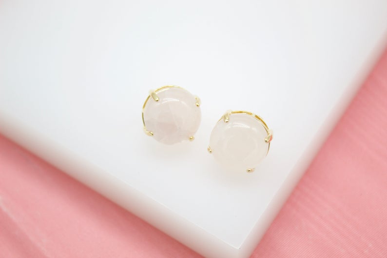 A pair of round rose quartz stud earrings featuring polished pink stones set in gold prongs rests on a white display surface.