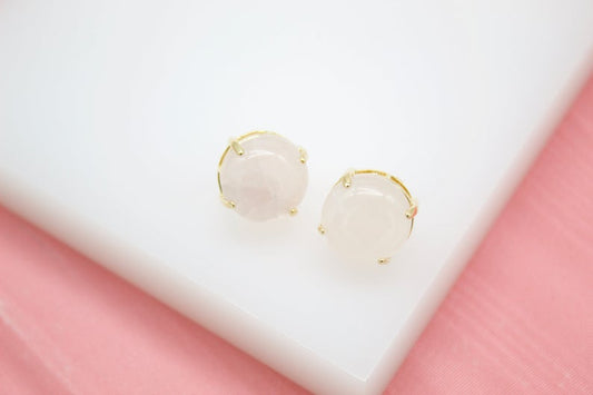 A pair of round rose quartz stud earrings featuring polished pink stones set in gold prongs rests on a white display surface.