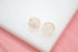 A pair of round rose quartz stud earrings featuring polished pink stones set in gold prongs rests on a white display surface.