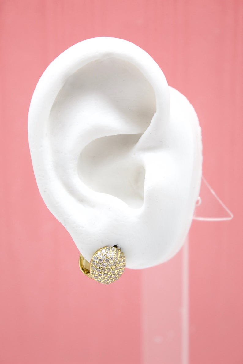 A white mannequin ear displays a sparkling gold pave huggie hoop earring against a pink background.