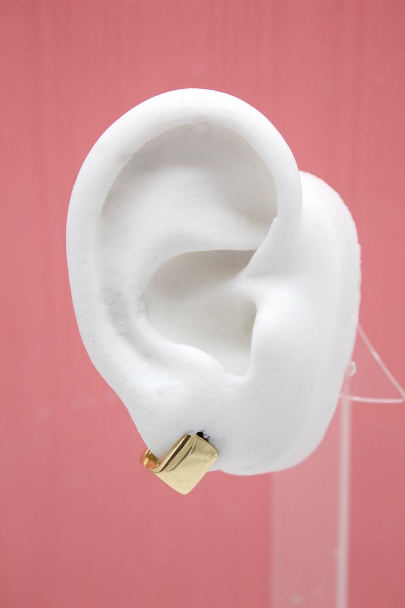 A white display ear model showcases a small, shiny gold huggie hoop earring against a pink background.