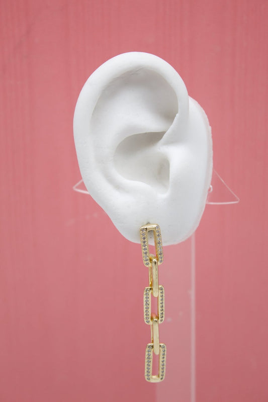A white mannequin ear displays a dangling gold chain link earring encrusted with sparkling cubic zirconia stones against a pink background.