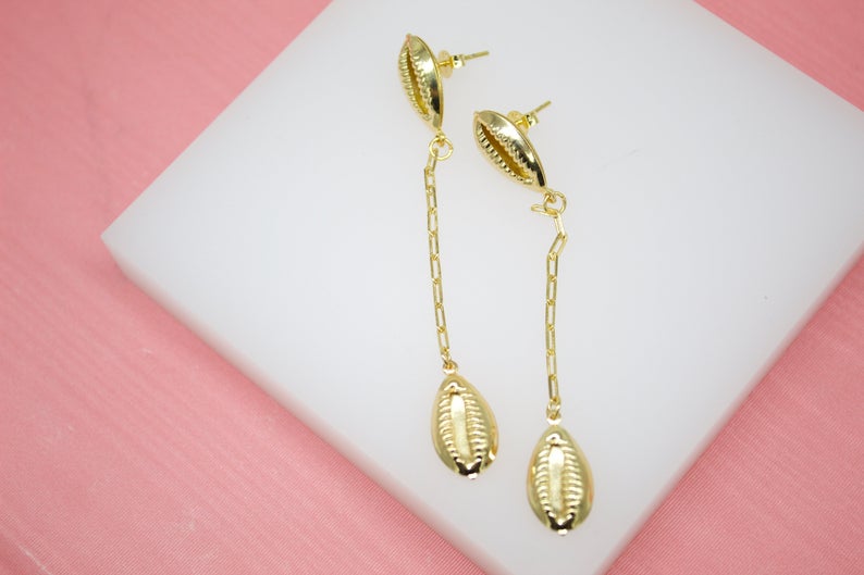 A pair of gold-plated cowrie shell drop earrings rests on a white display block against a pink background.
