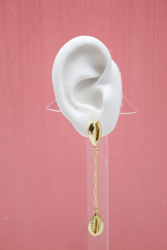 A white mannequin ear displays a dangling gold cowrie shell chain earring against a pink background.