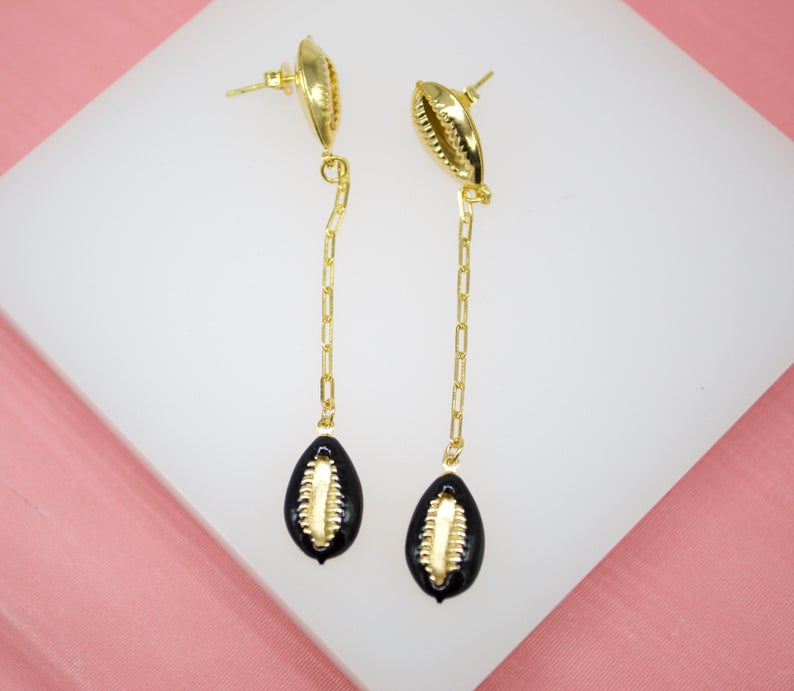 A pair of gold dangle earrings features cowrie shell studs connected by delicate chains to black and gold cowrie shell pendants.