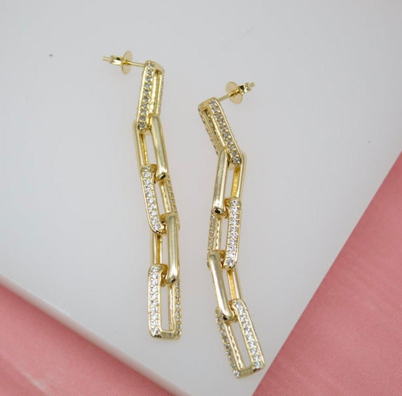 A pair of dangling gold chain-link earrings encrusted with sparkling pave diamonds lie on a white surface against a pink background.