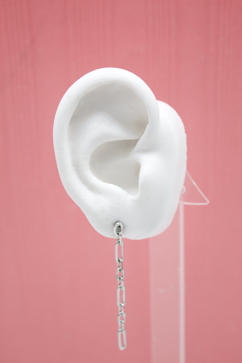 A white model ear on a pink background displays a dangling silver chain link earring.
