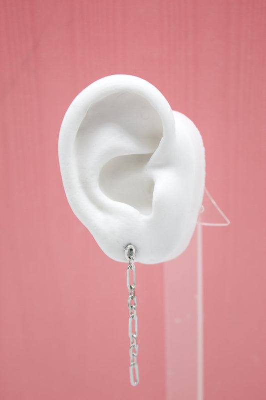 A white model ear on a pink background displays a dangling silver chain link earring.