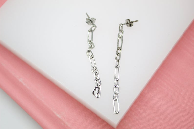 A pair of silver paperclip chain drop earrings rests against a white and pink display surface.