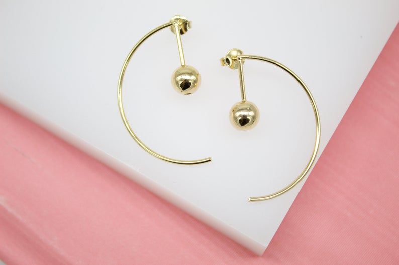 A pair of modern gold hoop earrings featuring a suspended ball design rests on a white display surface.