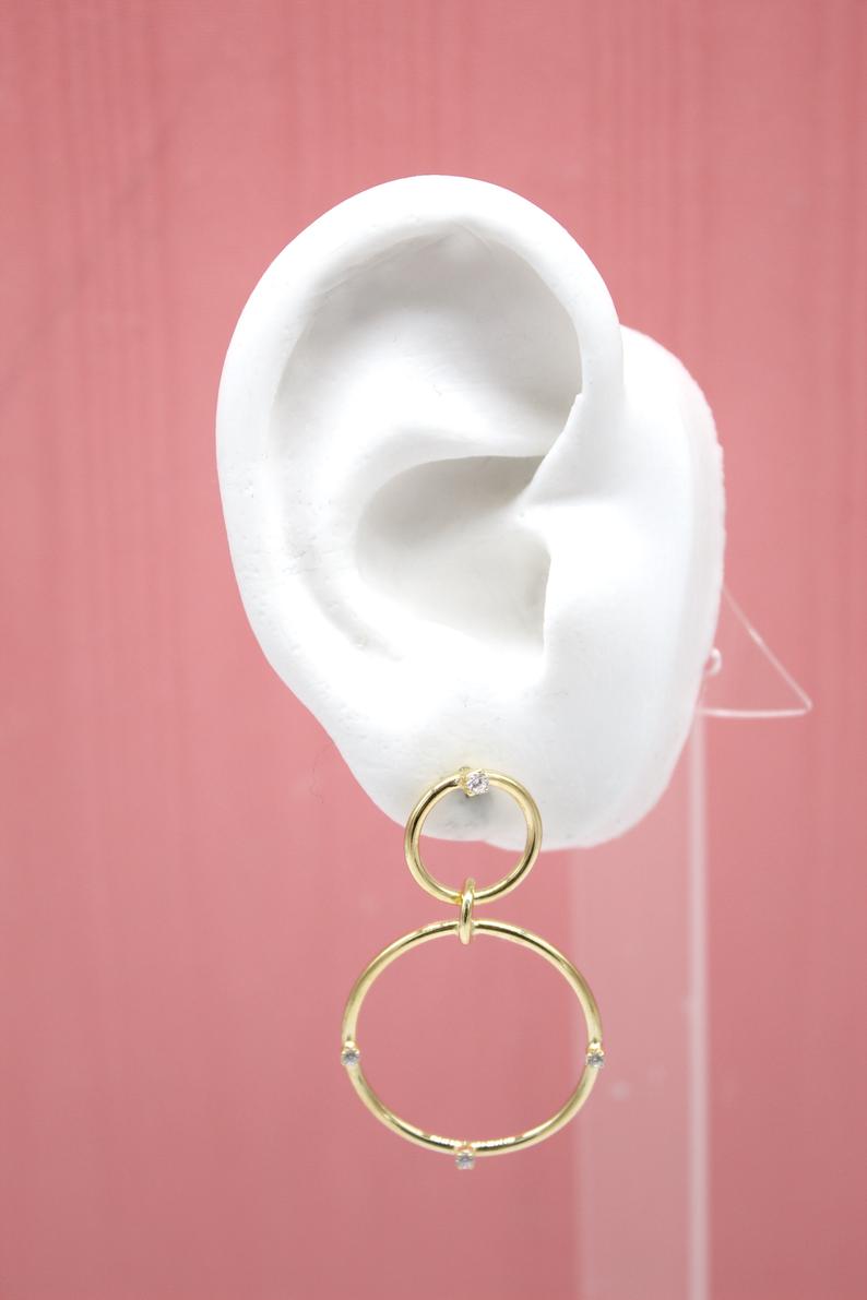A white mannequin ear displays a pair of elegant gold double-hoop earrings embellished with sparkling cubic zirconia stones against a pink background.