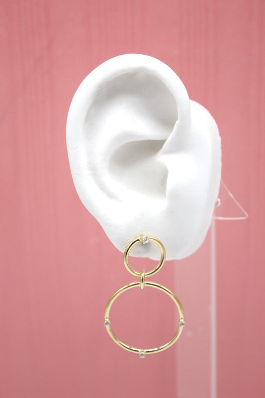 A white mannequin ear displays a pair of elegant gold double-hoop earrings embellished with sparkling cubic zirconia stones against a pink background.