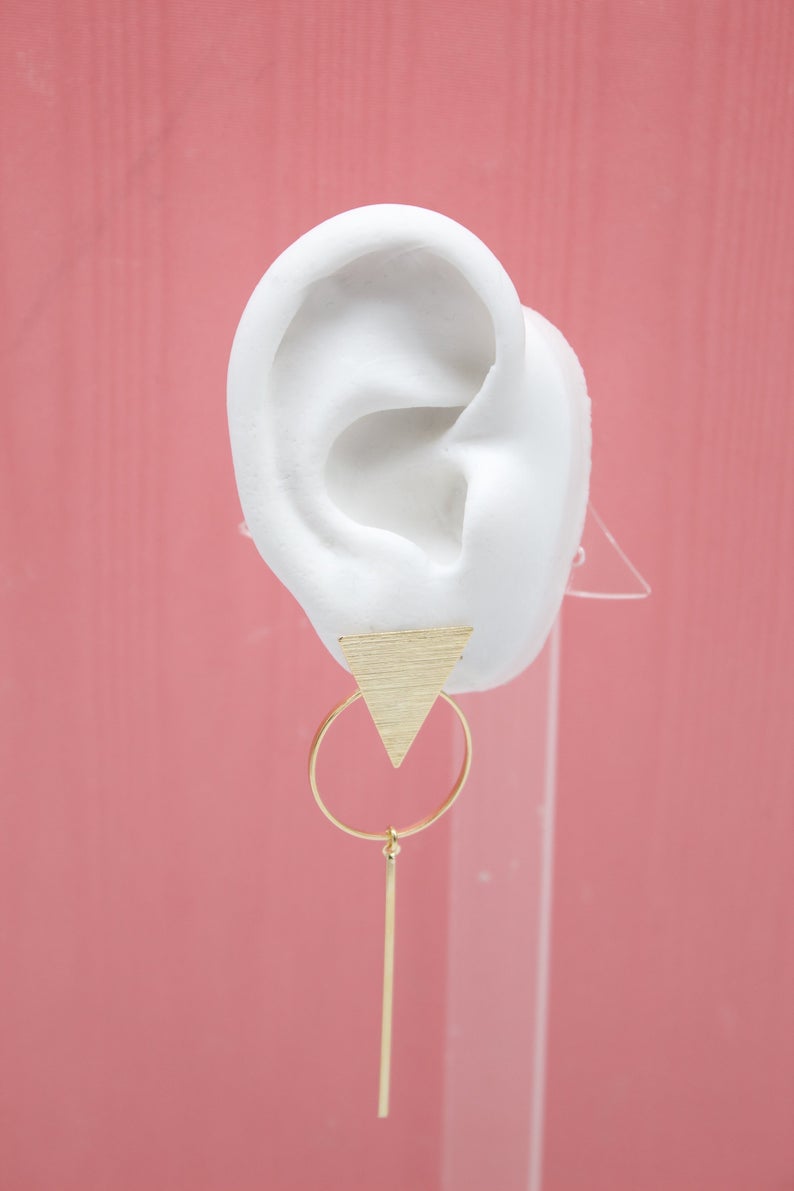 A white mannequin ear displays a modern gold geometric drop earring featuring a brushed triangle, a hoop, and a delicate bar.