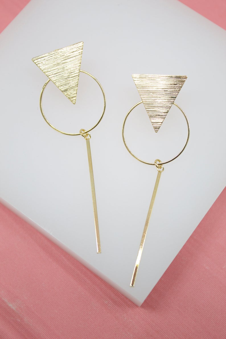 A pair of gold geometric drop earrings featuring a textured triangle, circle, and vertical bar display against a white and pink background.