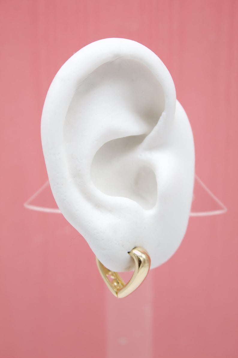 A white mannequin ear displays a polished gold heart-shaped hoop earring against a soft pink background.