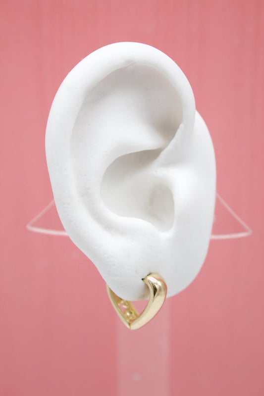 A white mannequin ear displays a polished gold heart-shaped hoop earring against a soft pink background.