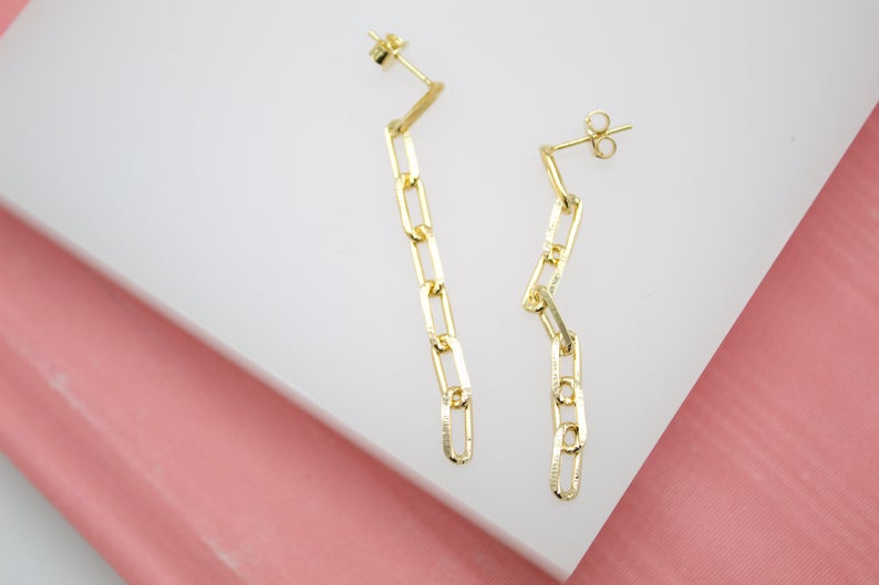 A pair of elegant gold chain link drop earrings rests against a white and pink display surface.