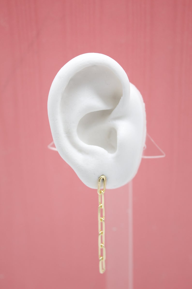 A white mannequin ear displays a dangling gold paperclip chain earring against a pink background.