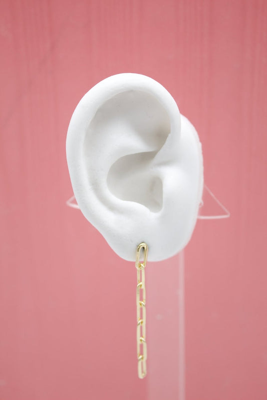 A white mannequin ear displays a dangling gold paperclip chain earring against a pink background.